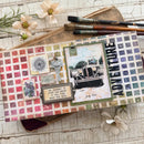 A decorative mixed-media journal or scrapbook box laid flat on a whitewashed wood surface, surrounded by flowers, paintbrushes, and patterned paper. The item features a grid of vintage-style stamps, collage elements, a key, a small plaque with text, and large vertical 'ADVENTURE' lettering.