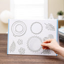 A hand is holding a clear acrylic embosser plate with various metal die-cut templates, including a fern, a flower, a sunburst, and a heart, set against a blurred indoor background.