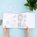 A person's hands hold open a white booklet showing metal die-cut craft templates, photographed from above on a light blue background with a green plant in the corner.
