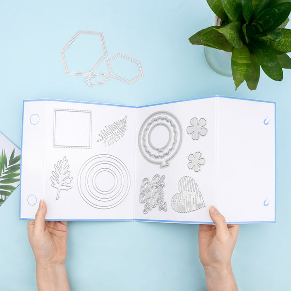 A person's hands hold open a white booklet showing metal die-cut craft templates, photographed from above on a light blue background with a green plant in the corner.