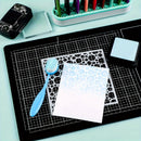 A crafting setup on a light blue surface featuring a black gridded cutting mat with a white square sheet, a blue-handled brush, a metal stencil, and a small black ink pad. A mint green marker tray holding colorful markers is visible in the background, along with a black decorative block and a clear acrylic block.