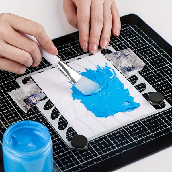 A close-up of hands mixing bright blue gel or paint on a white sheet over a black grid cutting mat, with a small blue jar in the foreground.