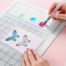 A close-up of a person using a white grid drawing tablet or coloring pad with a clear pen, while a butterfly-shaped sheet with printed butterfly designs lies on the surface. The scene is set against a soft pink background.