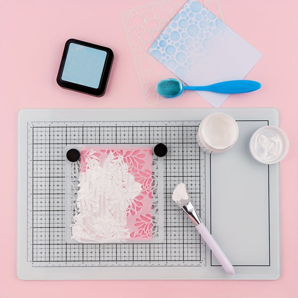 A flat-lay of a craft stamping kit on a pink background, featuring a white floral stamp on a pink block, a gray gridded cutting mat, a blue ink pad, a blue brush, and small white containers of wax or paste.