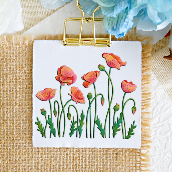 A square white card or small sign clipped at the top with a gold binder clip, featuring embroidered poppy flowers in pink, orange, and green on a burlap surface with decorative fabric and flowers around it.