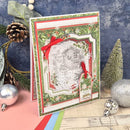 A handmade Christmas greeting card with a festive green and red holly border, a central white house illustration, and red ribbon accents, styled on a wooden surface with holiday decorations.