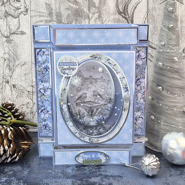 A decorative layered acrylic shadow box card featuring a winter Christmas theme, with blue and silver floral borders, a central oval window showing birds and snow, and a small 'Peace & Joy Christmas' plaque at the bottom. The card is styled on a gray surface with holiday ornaments, a pinecone, and a textured gray background.