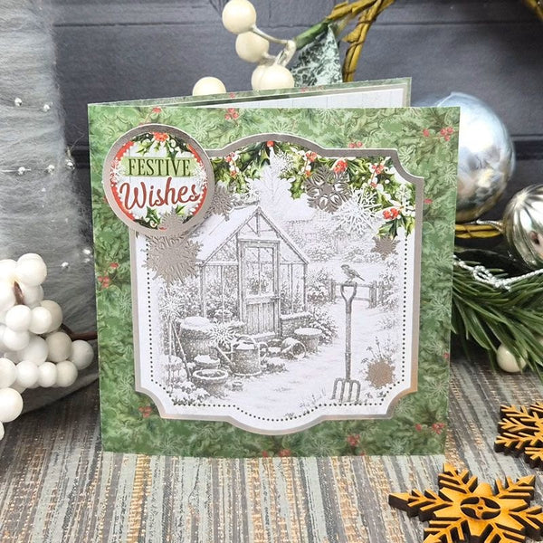 A handmade Christmas greeting card featuring a green patterned border, a central black-and-white illustration of a garden shed and bird feeder, and a round 'FESTIVE Wishes' embellishment. The card is styled on a rustic wooden surface with holiday decorations including white berries, pine branches, a silver ornament, and wooden snowflake ornaments.
