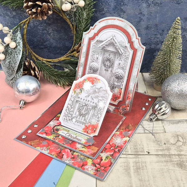A handmade pop-up Christmas card set displayed on a rustic wood surface with holiday decorations, including a pine wreath, silver ornaments, and a small tree. The cards feature a large arched card with a snowy house scene in black and white, layered over red floral and poinsettia-patterned cards with a smaller matching card standing in front.