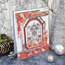 A handmade Christmas greeting card featuring a snowy front door scene surrounded by red poinsettias, displayed on a rustic gray surface with pinecones and a silver Christmas tree in the background.