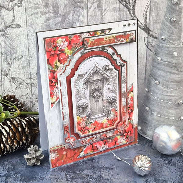 A handmade Christmas greeting card featuring a snowy front door scene surrounded by red poinsettias, displayed on a rustic gray surface with pinecones and a silver Christmas tree in the background.