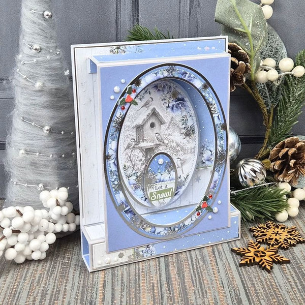 A layered square greeting card or decorative gift box with a winter village scene in the center, styled on a rustic surface with holiday greenery, pinecones, white berries, and silver ornaments.