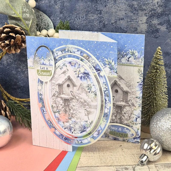 A handmade Christmas greeting card featuring a winter birdhouse scene, styled with holiday decorations including pinecones, ornaments, and a glittery tree against a blue textured backdrop.