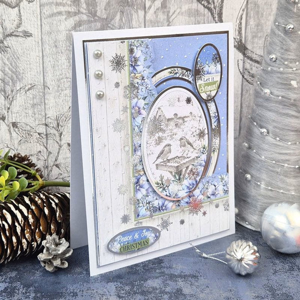 A handmade greeting card with a winter Christmas theme, featuring a snowy oval window, blue floral accents, and 'Peace & Joy Christmas' text, styled on a rustic gray surface with pinecones and holiday decorations.