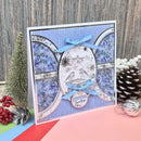A handmade greeting card with a blue and white striped background, floral arches, a central oval with two birds, and light blue ribbon bows, styled with holiday props on a pink surface.