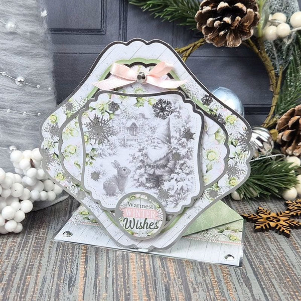 A decorative handmade greeting card with layered scalloped edges, a pale pink bow, and a central winter scene featuring a gnome, squirrel, snowflakes, and the words 'Warmest Winter Wishes.' The card is styled on a rustic wood surface with pine branches, pinecones, white berries, and holiday ornaments in the background.