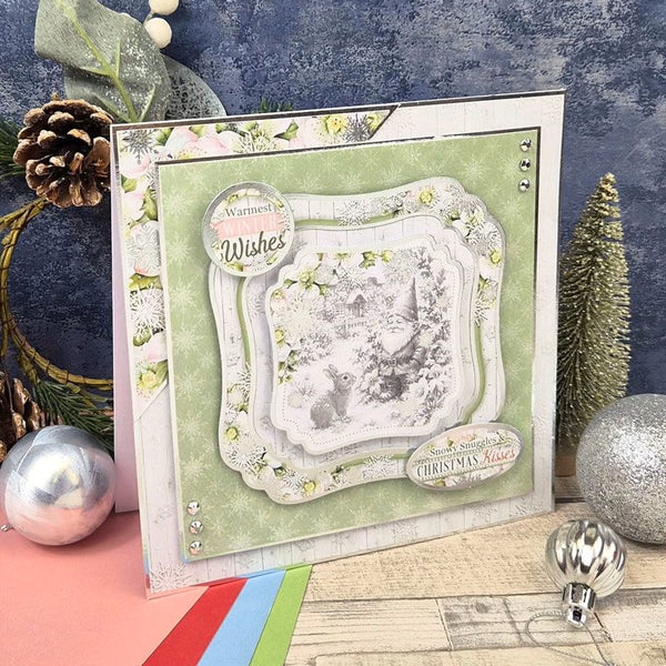 A handmade layered Christmas greeting card with a light green background, white ornate borders, and a central winter scene, styled on a tabletop with holiday ornaments and a blue textured backdrop.