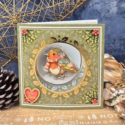 A square handmade greeting card featuring a detailed illustration of a robin bird wearing a red and cream scarf, set against a winter wreath design. The card is styled with a pinecone, dried botanicals, and a blue textured background.