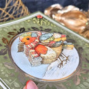 A close-up of a decorative card featuring a 3D layered robin bird wearing a red and white striped scarf, set against a green holly-patterned background.