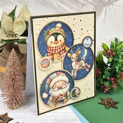A handmade Christmas greeting card featuring three circular illustrations: a penguin in a winter hat and scarf, a reindeer under a full moon, and Santa Claus. The card is styled on a white wooden surface with holiday decorations, including a small glittery Christmas tree, poinsettias, and greenery.