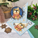 A handmade Christmas card featuring a penguin wearing a yellow beanie and red-and-white plaid scarf, displayed on a light blue snowflake-patterned card. The card is styled with holiday decorations including a miniature pine tree, burlap pot, and green foliage on a white wood surface.