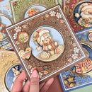 A hand holds a square decorative card featuring a teddy bear illustration in a winter outfit, surrounded by ornate floral and scroll borders. Other similar cards are partially visible in the background.