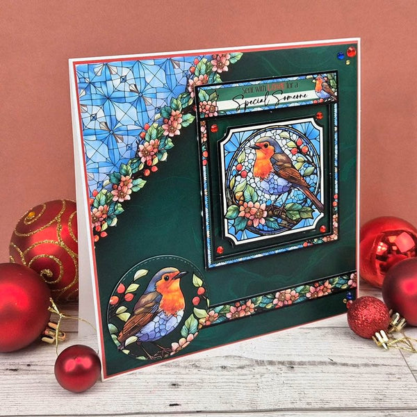 A handmade Christmas card featuring robin birds in stained-glass style, displayed on a rustic white wooden surface with red Christmas ornaments against a terracotta background.