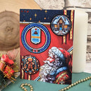 A festive Christmas card featuring Santa Claus and stained-glass-style holiday illustrations, styled on a tabletop with a candle, wooden slice, beads, and small holiday decor.