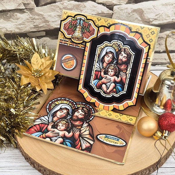 A set of Christmas nativity cards displayed on a rustic wood slice with holiday decorations, including gold tinsel, a gold poinsettia, a gold bell, and red and gold ornaments. The cards feature colorful illustrations of the Holy Family (Mary, Joseph, and baby Jesus) in a stained-glass style.