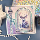 A square decorative card featuring a painted deer in a winter scene, surrounded by layered arch frames and ornate borders. The card has a lavender and teal color palette with glitter accents, photographed on a blue surface with decorative props including a pearl strand, a white star-shaped object, and holographic paper in the background.