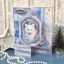 A handmade layered card featuring a white fox in a snowy blue and purple winter design, displayed alongside a small silver star decoration, a miniature tree, and white beads on a blue surface.