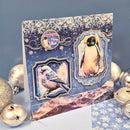 A handmade penguin-themed greeting card with layered blue and silver glitter, penguin illustrations, and decorative bird frames, styled with silver Christmas ornaments on a blue surface.