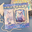 A decorative owl-themed card or art piece displayed on a blue surface, featuring two owl illustrations, ornate blue and gold borders, rhinestone accents, and a small matching tag. A pearl bead strand, envelope, and iridescent paper are arranged around it as styling props.