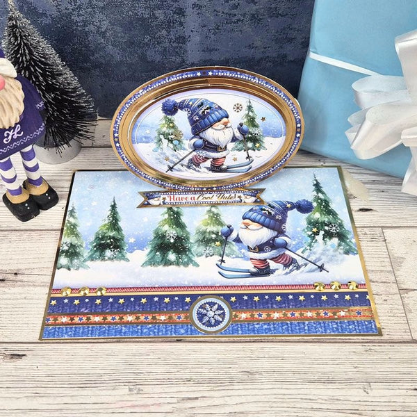 A holiday-themed kitchen or dining set displayed on a light wood surface, featuring a rectangular placemat and an oval platter with a gnome skiing among snowy trees. The set is styled with a small decorative tree, a gift box, and a nutcracker figurine.