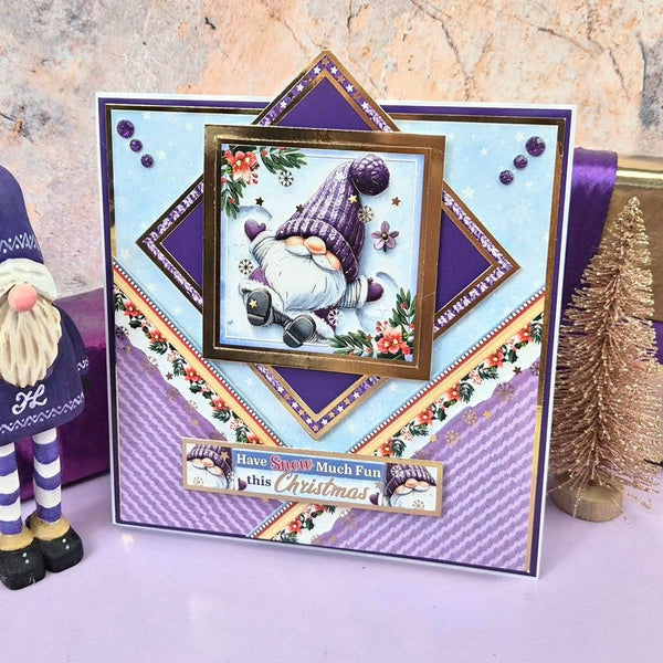 A handmade Christmas gnome greeting card displayed on a lavender surface, featuring a purple and light blue geometric design with a central framed illustration of a sleeping gnome. A small decorative gnome figurine and a gold glitter tree stand beside the card against a textured stone background.
