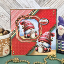 A square handmade Christmas greeting card featuring two festive gnomes in red knit hats and blue beards, surrounded by holiday decorations including a gingerbread house cup, gold bead garland, and a gnome figurine, set against a rustic whitewashed wood background.