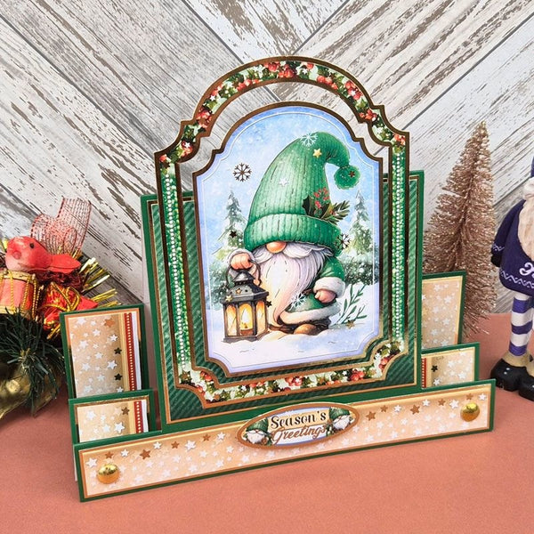 A decorative Christmas advent-style box with a central illustrated gnome in a green hat holding a lantern, surrounded by green and cream panels with gold stars and red berry accents. The front reads "Season's Greetings.