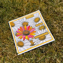 A square printed card or art print featuring illustrated daisies and a bright pink-orange-yellow flower, placed on grass. The print has yellow border lines and small text boxes reading phrases like 'Blossom wildly' and 'someone?'.