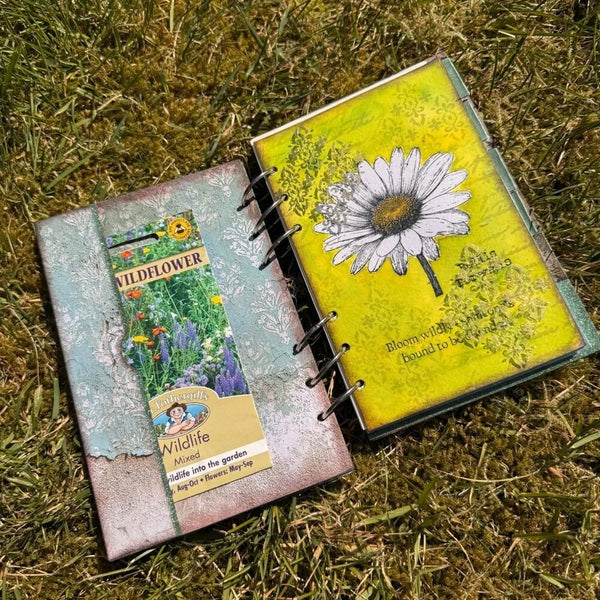 Two small spiral-bound art notebooks or journal pages laid on grass. One page has a distressed teal and pink cover with a seed packet graphic reading Wildflower and a Wildlife Mound label.