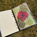 An open spiral-bound notebook lying on grass, showing lined paper on one side and a decorated page with a red poppy illustration and the word 'Papaver' on the other.