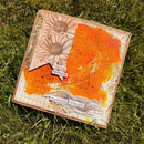 A square handmade art card or scrapbook board placed on green grass, featuring distressed orange and cream backgrounds, a daisy illustration, torn paper elements, and printed phrases.