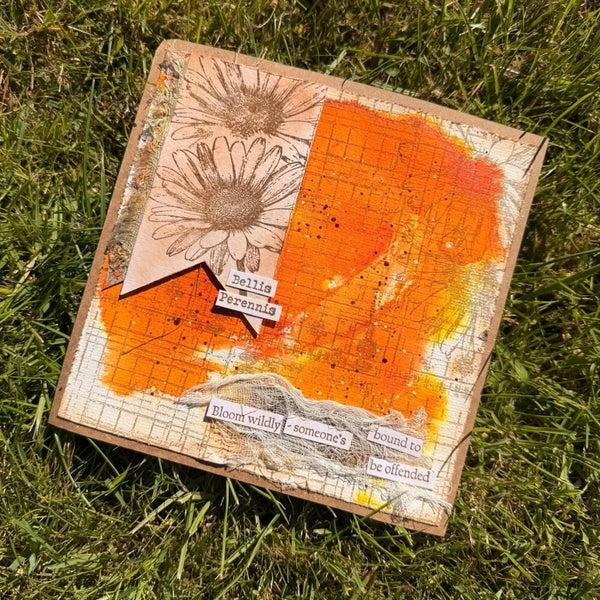 A square handmade art card or scrapbook board placed on green grass, featuring distressed orange and cream backgrounds, a daisy illustration, torn paper elements, and printed phrases.
