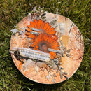 A round mixed-media art plate or tray decorated with two orange daisy-like flowers, textured paper, leaves, and small paper labels, placed on green grass outdoors.
