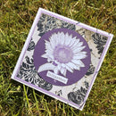 A handmade square greeting card featuring a white and purple dahlia flower on a purple circular background, set against a black and white damask pattern. The card is placed on green grass, with the word 'helianthus' written on a small banner.