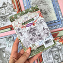 A hand holds a square scrapbook paper card featuring a snowy village scene and the title 'An Artist's Garden at Christmas' by Melted Memories. Other patterned paper sheets are visible in the background.