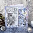A handmade winter-themed greeting card displayed on a rustic gray wooden surface with holiday decorations. The card features a central black-and-white illustration of a snow-covered mailbox, blue floral patterns, silver snowflakes, and a decorative oval label that reads 'Let it Snow!'.