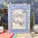 A handmade layered greeting card featuring a winter landscape with two ducks in the snow, surrounded by a blue floral border. The card is styled with holiday props including pinecones, a miniature tree, and silver ornaments against a textured blue background.