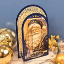 A Christmas-themed arched card or decorative plaque featuring an illustration of Santa Claus, styled on a tabletop with gold ornaments and a blue backdrop.