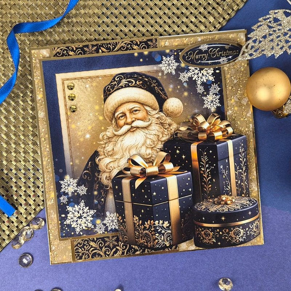 A square Christmas greeting card featuring Santa Claus with wrapped presents, surrounded by gold and navy decorative borders and holiday ornaments, photographed on a blue surface with ribbon and gold accents.